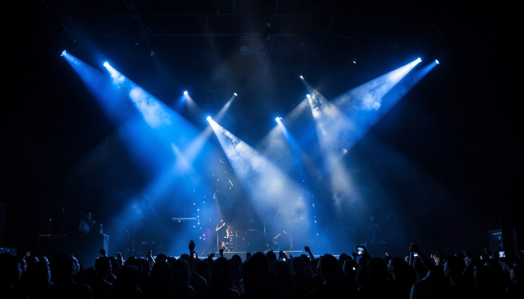Dark atmospheric stage with dramatic blue and white spotlight beams cutting through haze, evoking the energy of a K-pop concert performance