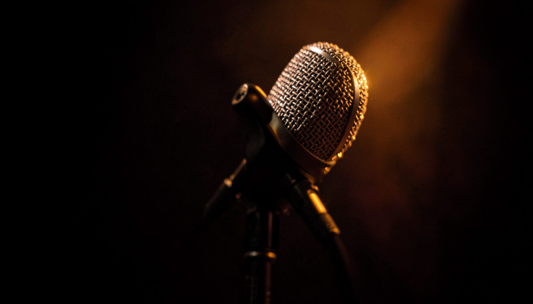 A close-up of a microphone on a dark stage with warm amber backlighting, suggesting an intimate broadcast setting