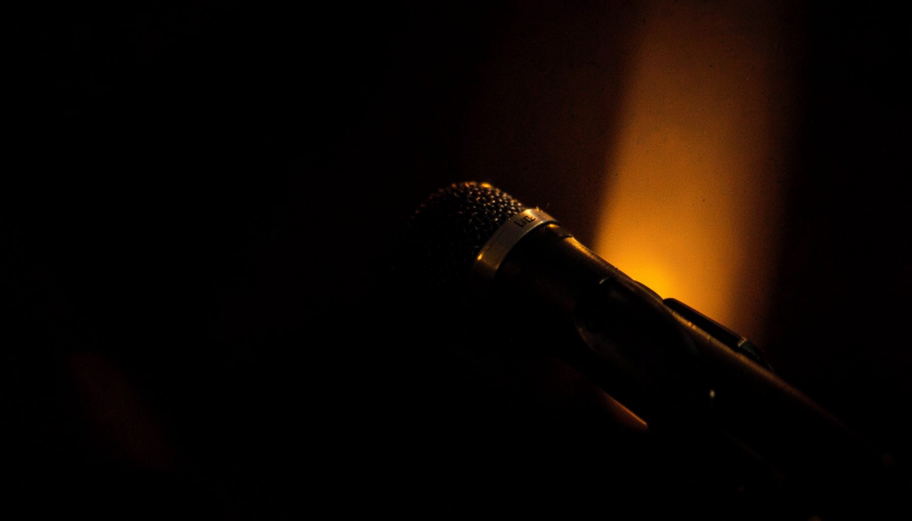 A close-up of a microphone on a dark stage with warm amber backlighting, suggesting an intimate interview setting