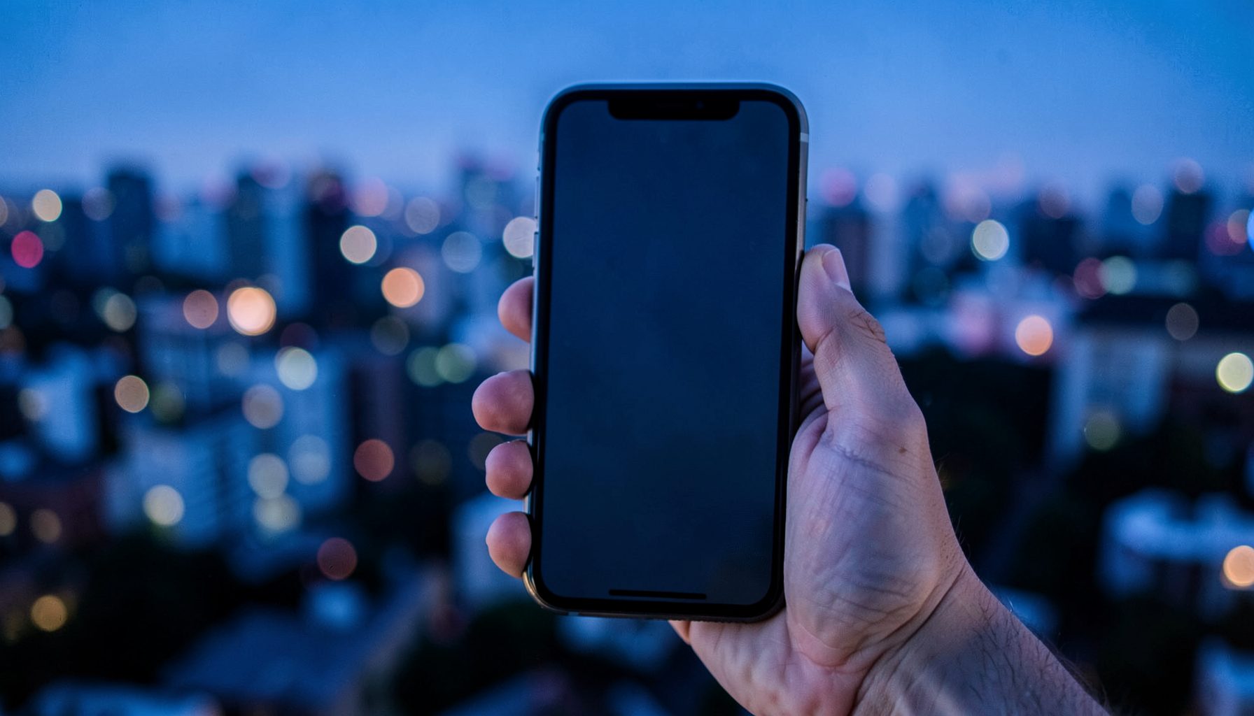 A smartphone held in a hand against a blurred nighttime city backdrop, cool blue and purple tones dominating
