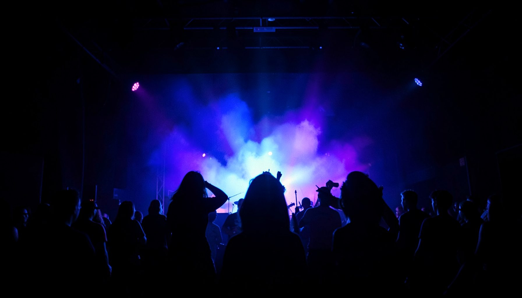 Dark stage with dramatic blue and purple concert lighting, silhouettes of performers against a glowing backdrop