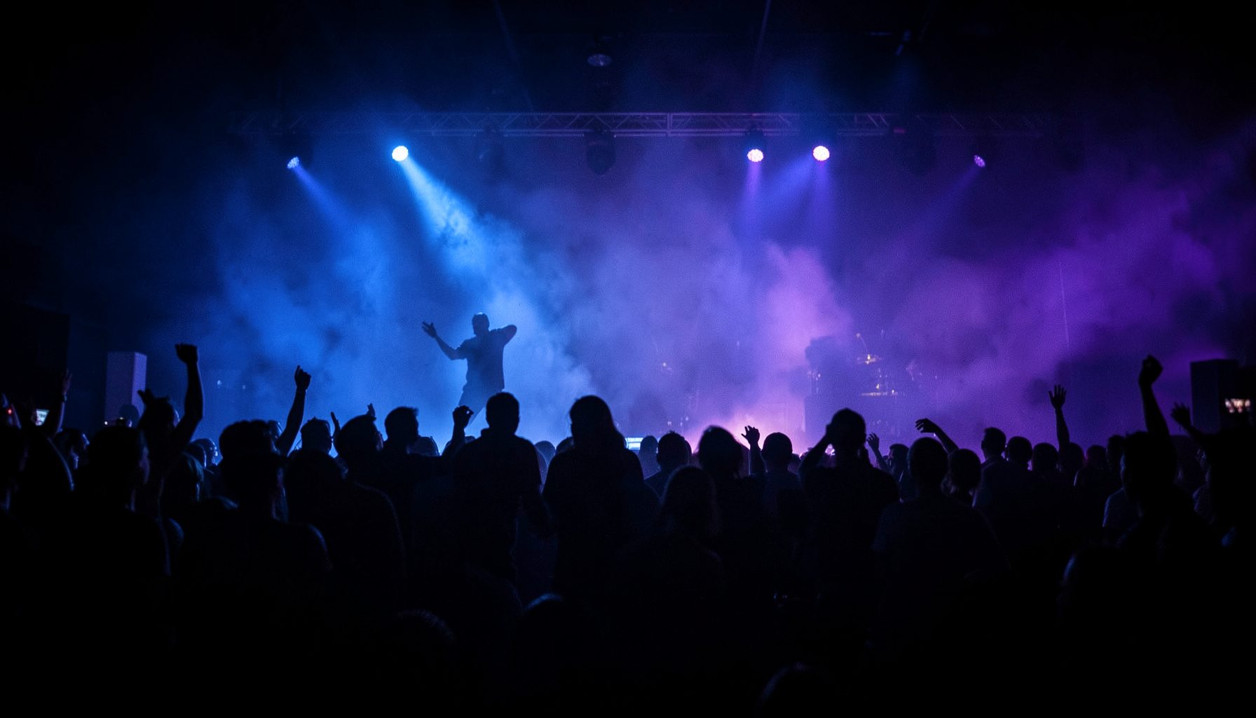 Dark stage with dramatic blue and purple lighting, silhouettes of performers visible through haze, concert atmosphere